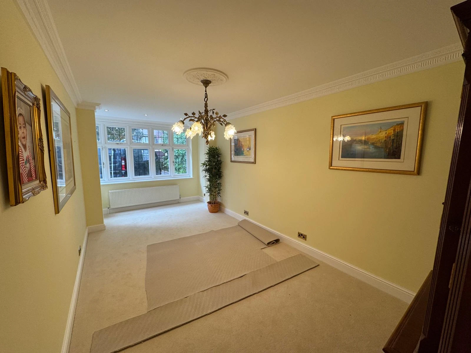 Spacious, bright room with light yellow walls, a decorative chandelier, framed artwork, and a plant. Carpet remnants are present.