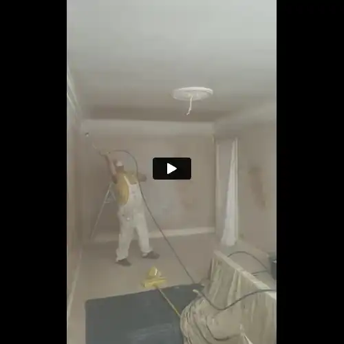 A person spray-painting the ceiling of a room while standing on a ladder, surrounded by draped furniture and paint tools.