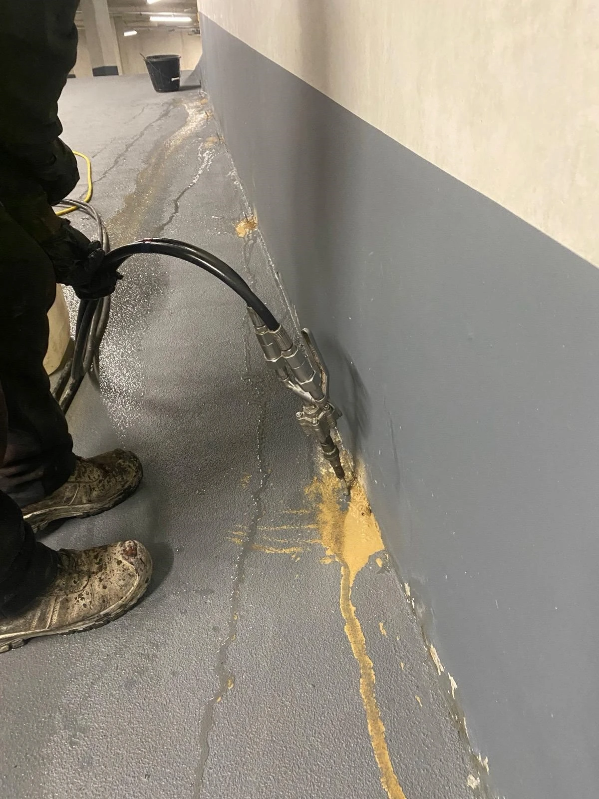 A worker operates a hose in a parking garage, applying a substance to a wall with visible leakage and grey floor.
