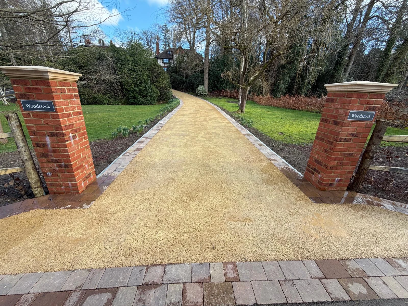 A pathway leading to a garden, framed by brick pillars labeled "Woodstock," with green grass and trees in the background.
