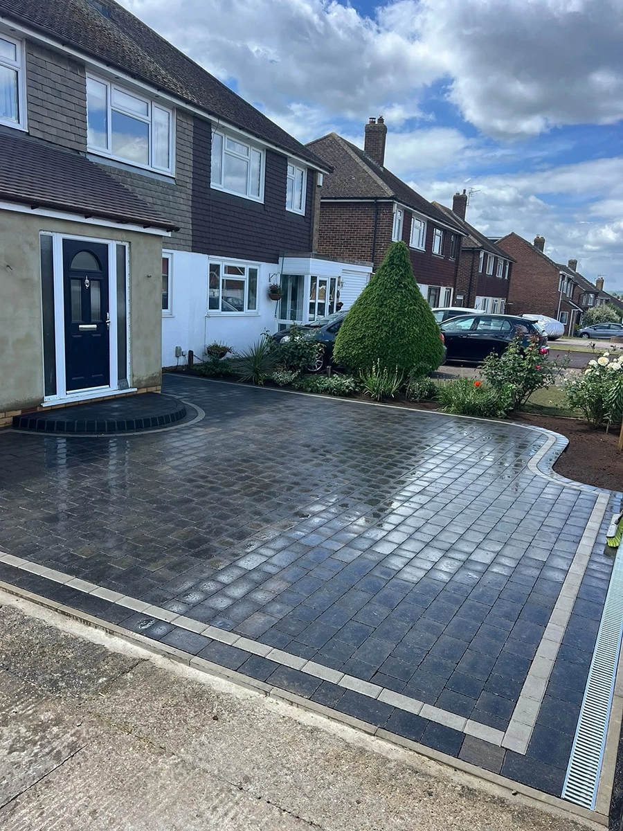Wet paved driveway in front of a two-story house with dark blue door and a triangular trimmed shrub near the driveway border, under a partly cloudy sky.