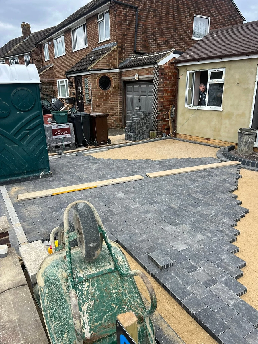 Residential driveway under construction with gray interlocking bricks partially laid on a sandy base, a green cement mixer in the foreground, wooden planks across the bricks, a man looking out from a window of a newly plastered house section, and a portable toilet and construction materials near a brick house in the background.