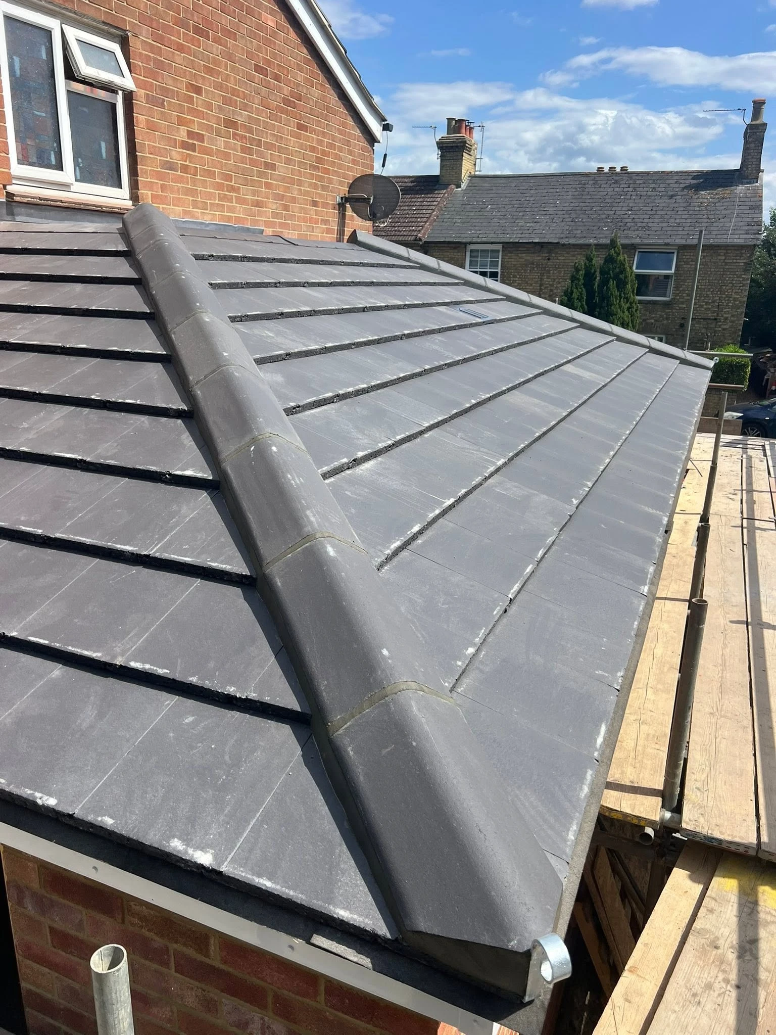 Newly tiled grey roof with ridge tiles, adjacent to a brick house and scaffolding on the right, under a partly cloudy blue sky.