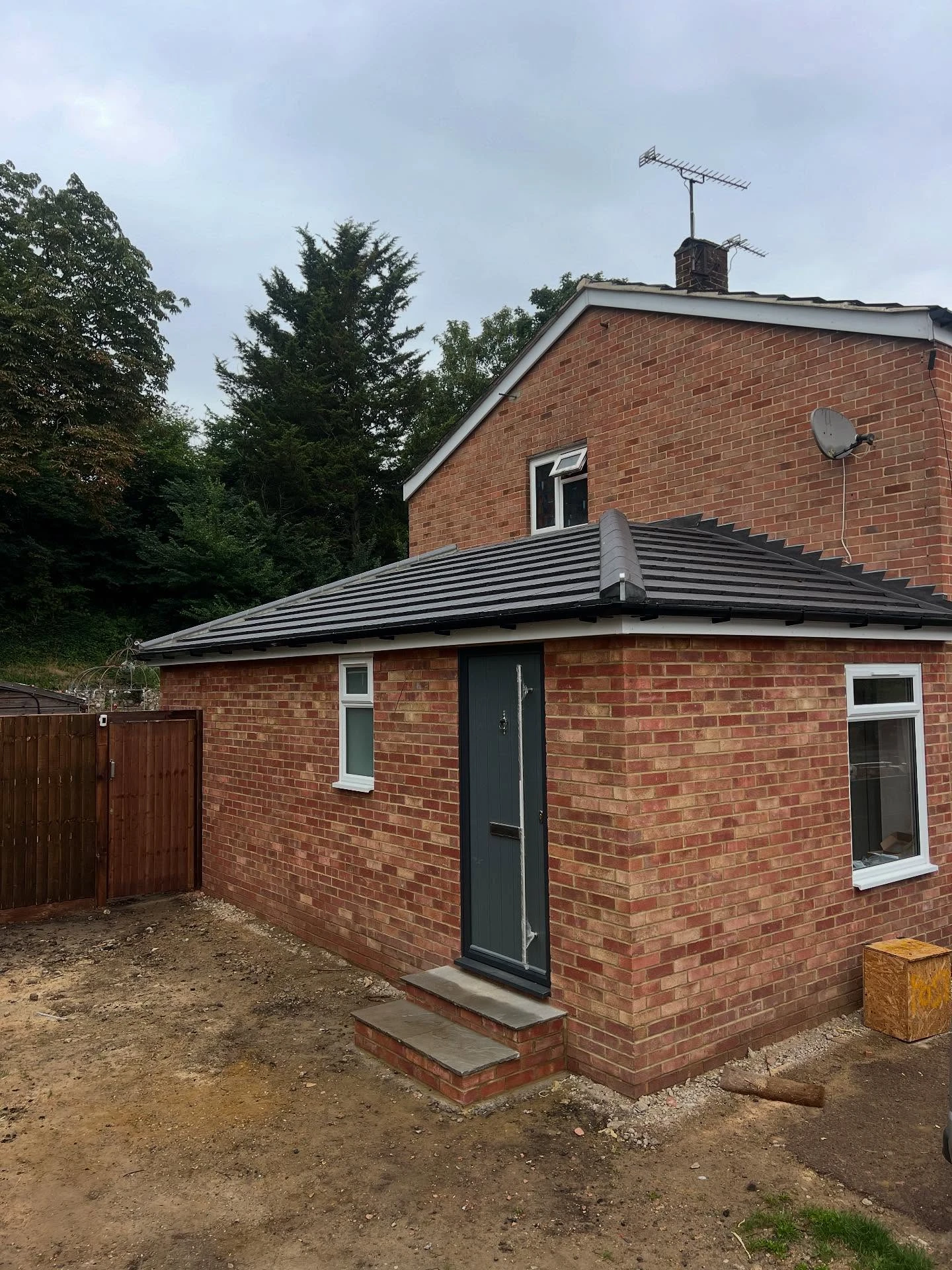 Newly constructed brick house extension with dark gray tiled roof, two small white-framed windows, a dark gray door with steps, a wooden fence gate, and trees in the background under a cloudy sky.