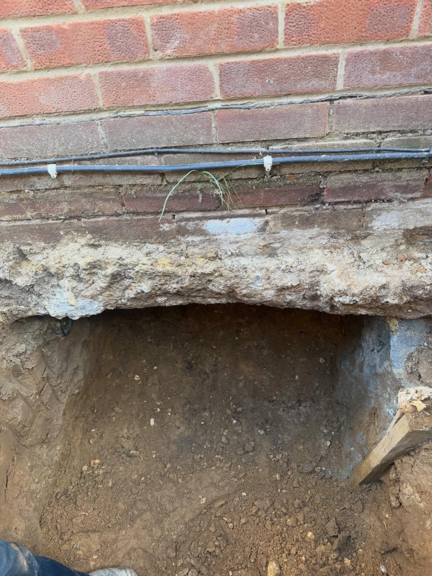 Excavated hole in front of a brick wall exposing soil and dirt with a patch of grass sprouting near the middle of the wall, and visible black cables secured with clips above the hole.