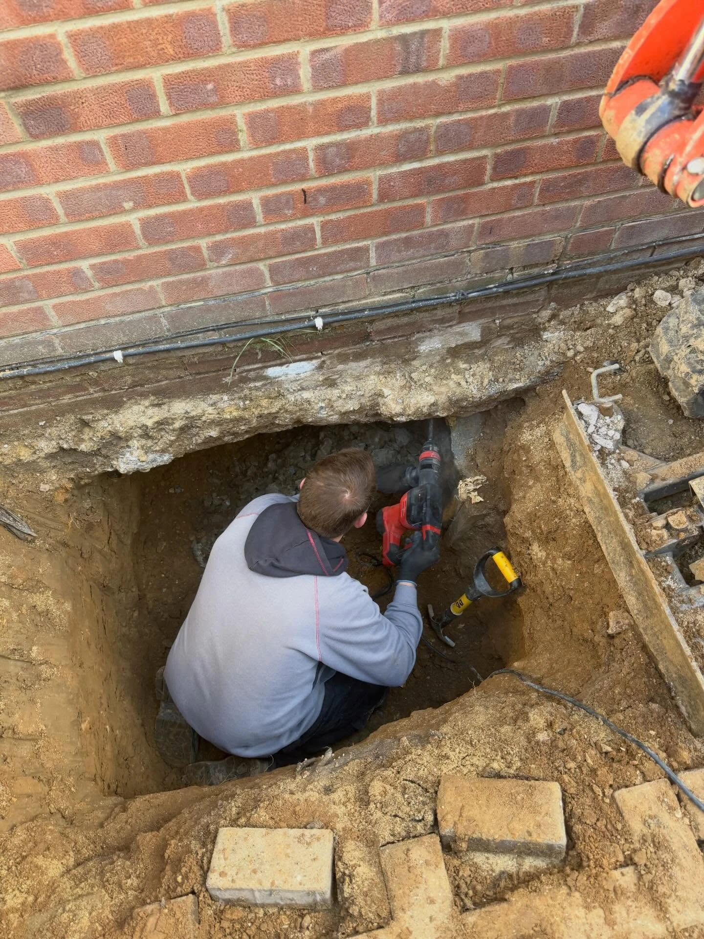 Man wearing gray hoodie and black gloves crouched in a deep rectangular hole dug next to a brick building wall, using a red power drill to work on the foundation area, with a metal tool and excavator arm visible nearby.