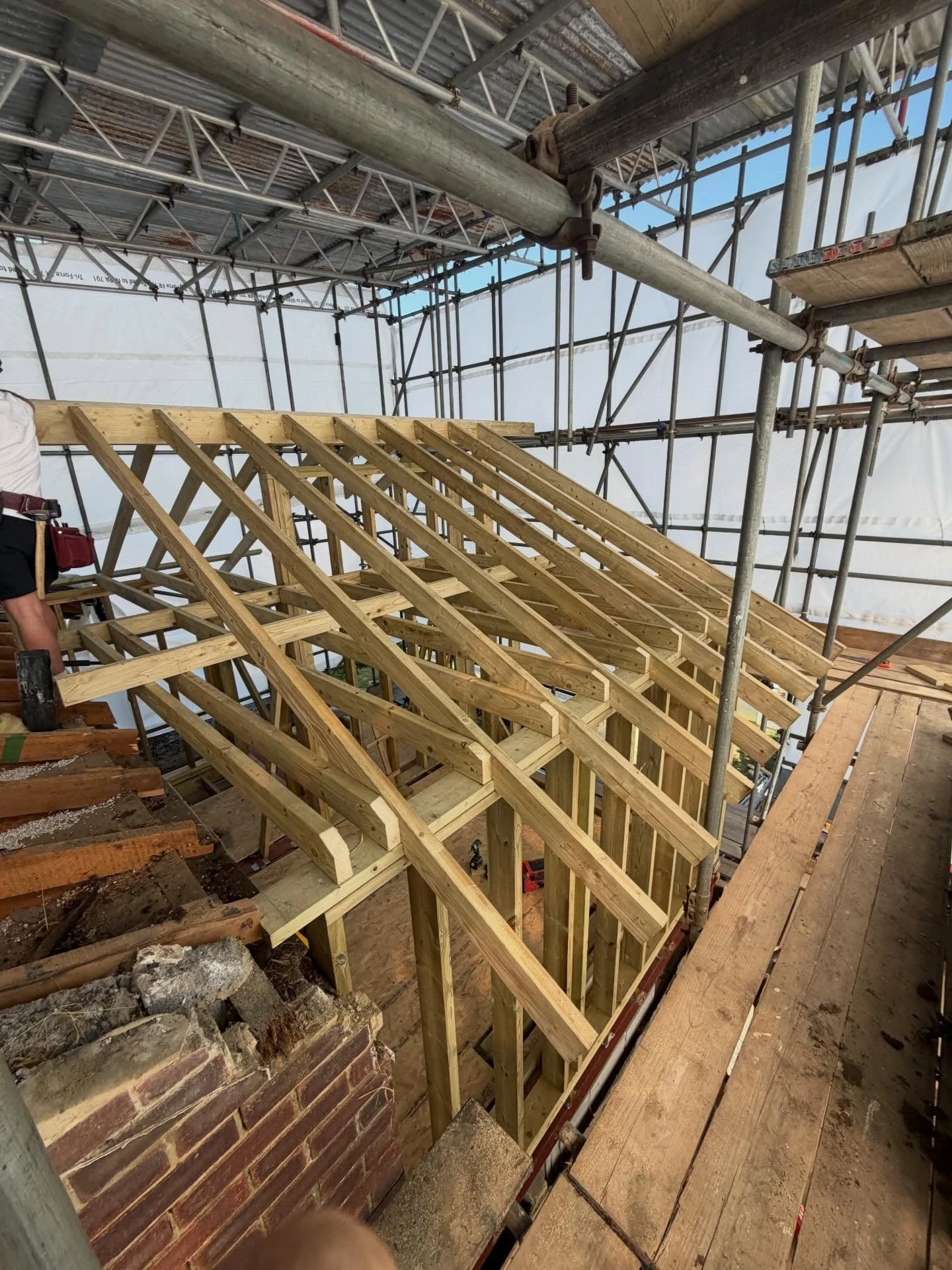 Framed wooden roof structure under construction surrounded by metal scaffolding with a partially visible worker on the left side.