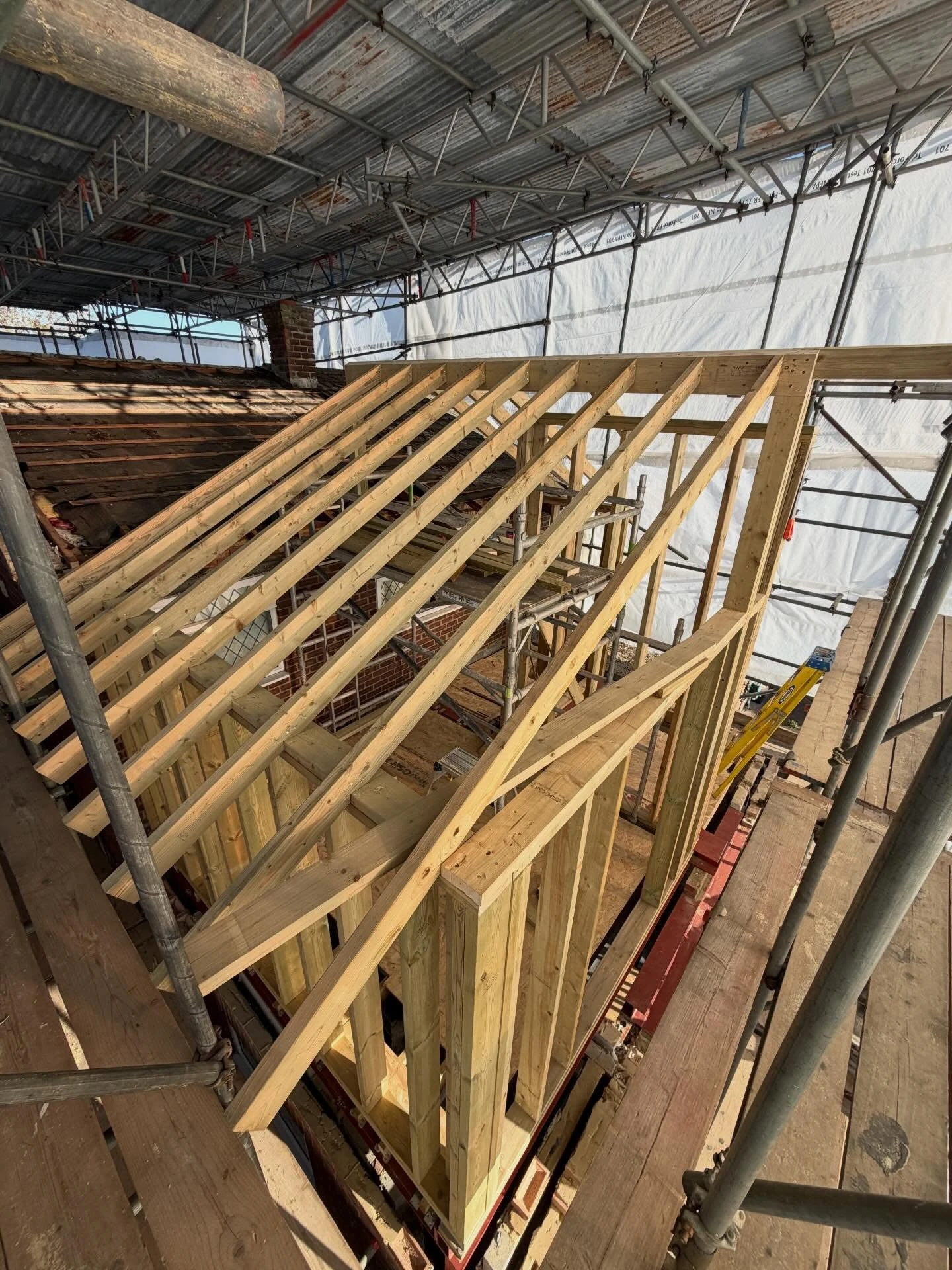 Wooden roof frame structure under construction surrounded by scaffolding inside a partially covered building, showing angled roof beams and vertical wall studs.
