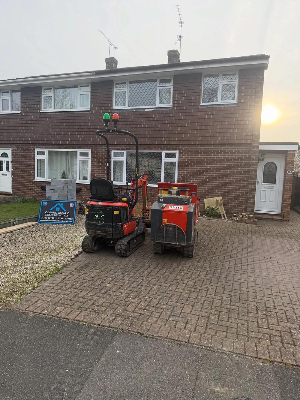 A semi-detached brick house with diamond-patterned windows and white doors, with construction equipment, including a small red digger and a red machine, parked on a brick driveway, and a blue Daniel Gould Construction sign with contact details on the left.
