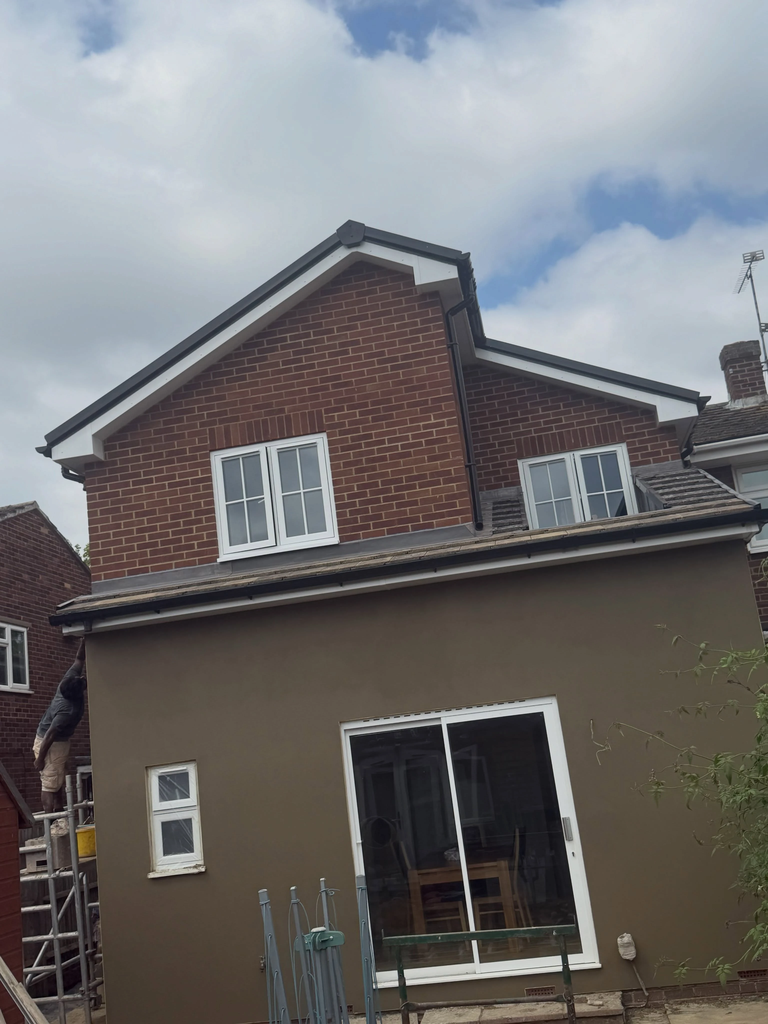 Rear view of a two-story house with a brown brick upper floor and a smooth brown lower extension, featuring two upper white-framed windows and a large sliding glass door below; a man is on scaffolding working on the side wall, with an overcast sky in the background.