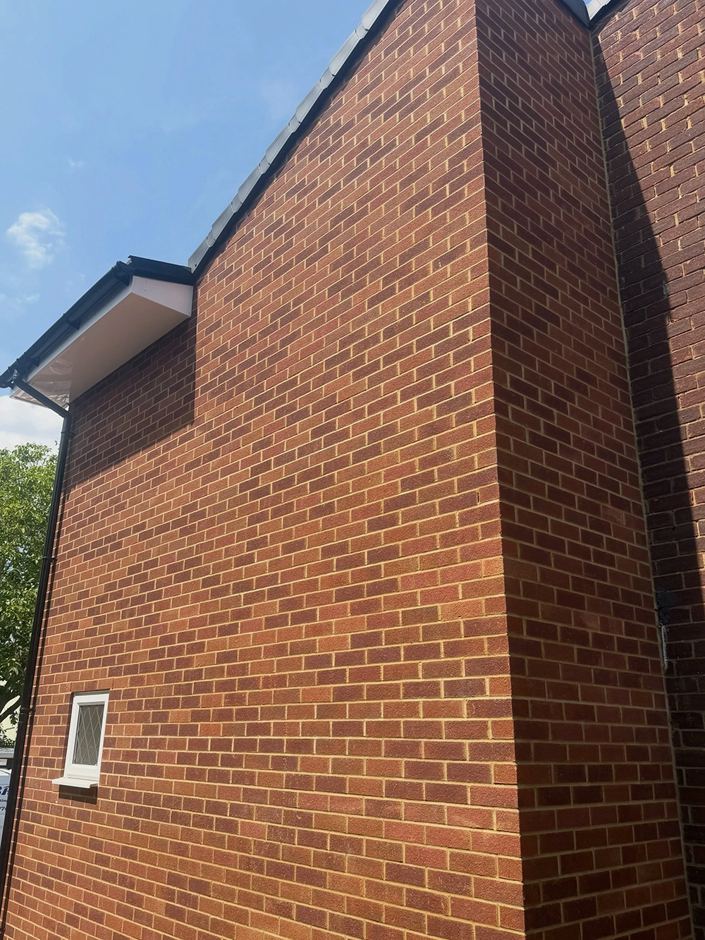 Close-up of a red brick building wall with a single small window with a white frame and diamond pattern glass, under a blue sky with some clouds, and part of a black gutter system on the left edge.