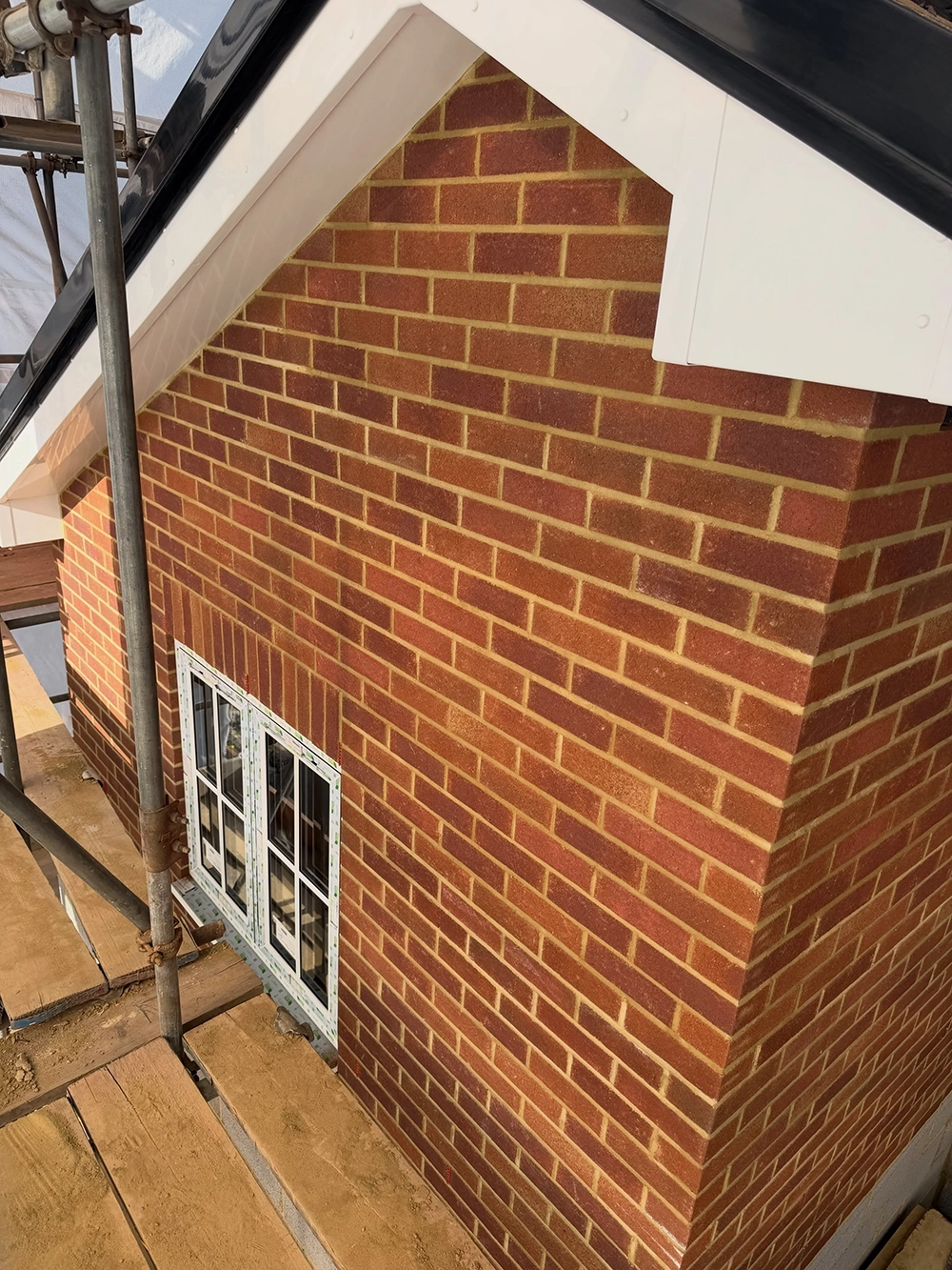 Newly constructed brick house corner with a white-framed window and scaffolding with wooden planks around it during construction.