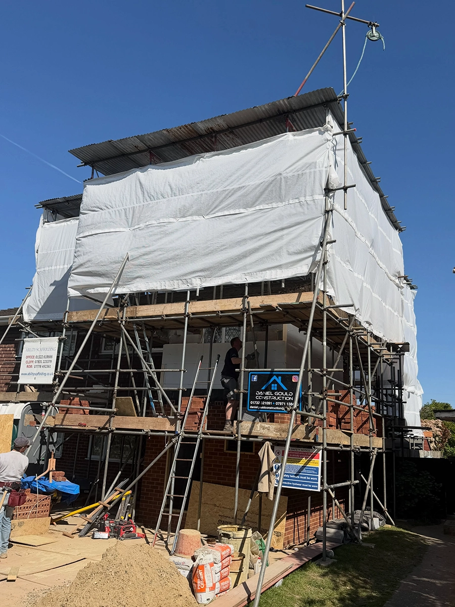 Two-story brick house under renovation with scaffolding and white protective sheeting covering the upper part, construction materials and tools on the ground, and a worker on the scaffolding.