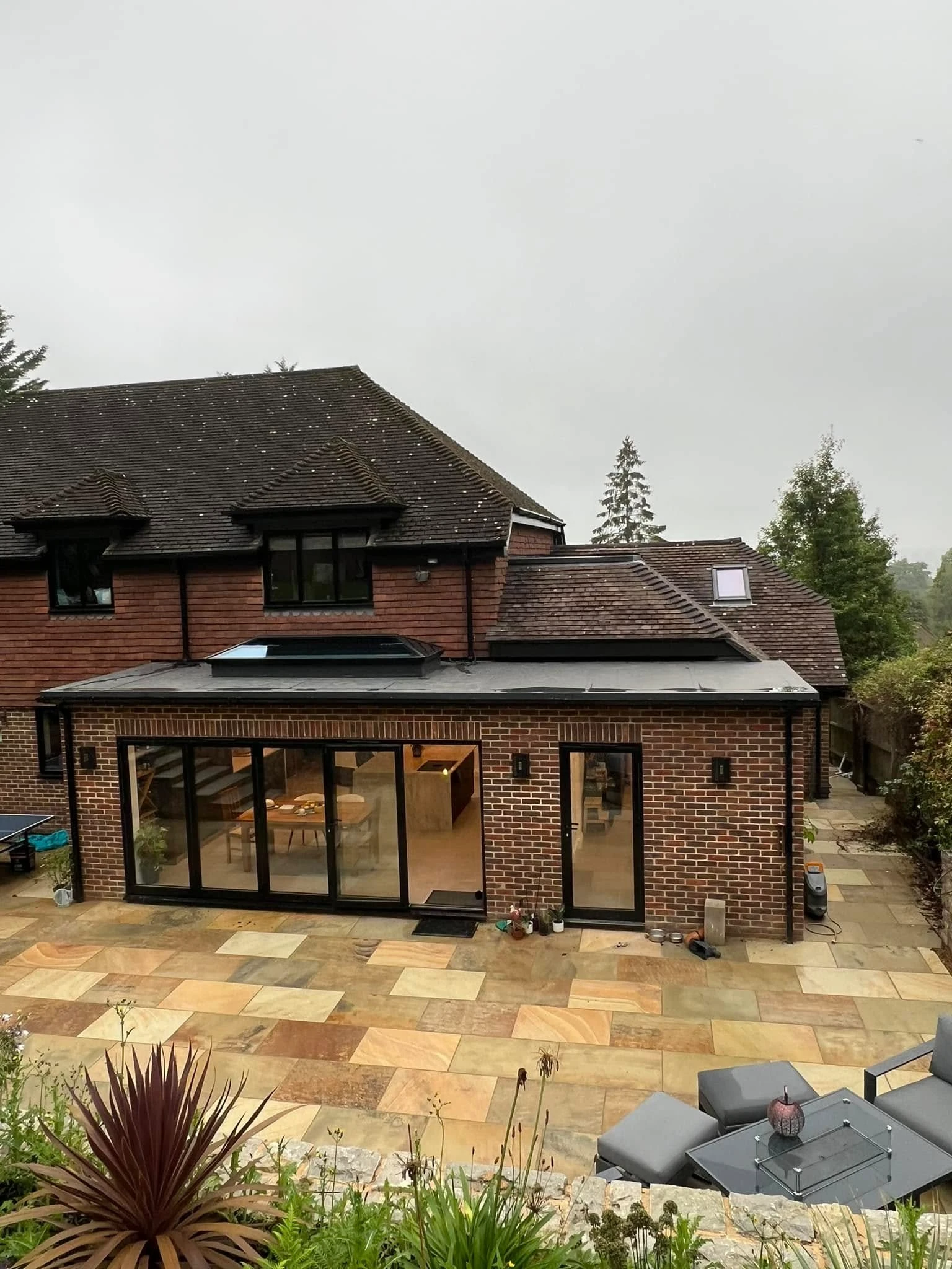 Back view of a two-story brick house with a modern single-story extension featuring large sliding glass doors and a single glass door, opening onto a paved patio with outdoor seating and planter in the foreground under an overcast sky.