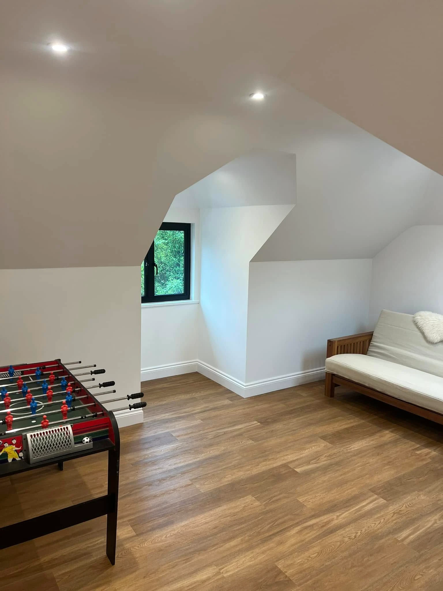 Bright room with wooden floor featuring a foosball table on the left and a white cushioned wooden sofa with a white fuzzy pillow on the right, under sloping white ceilings and a small black-framed window showing greenery outside.