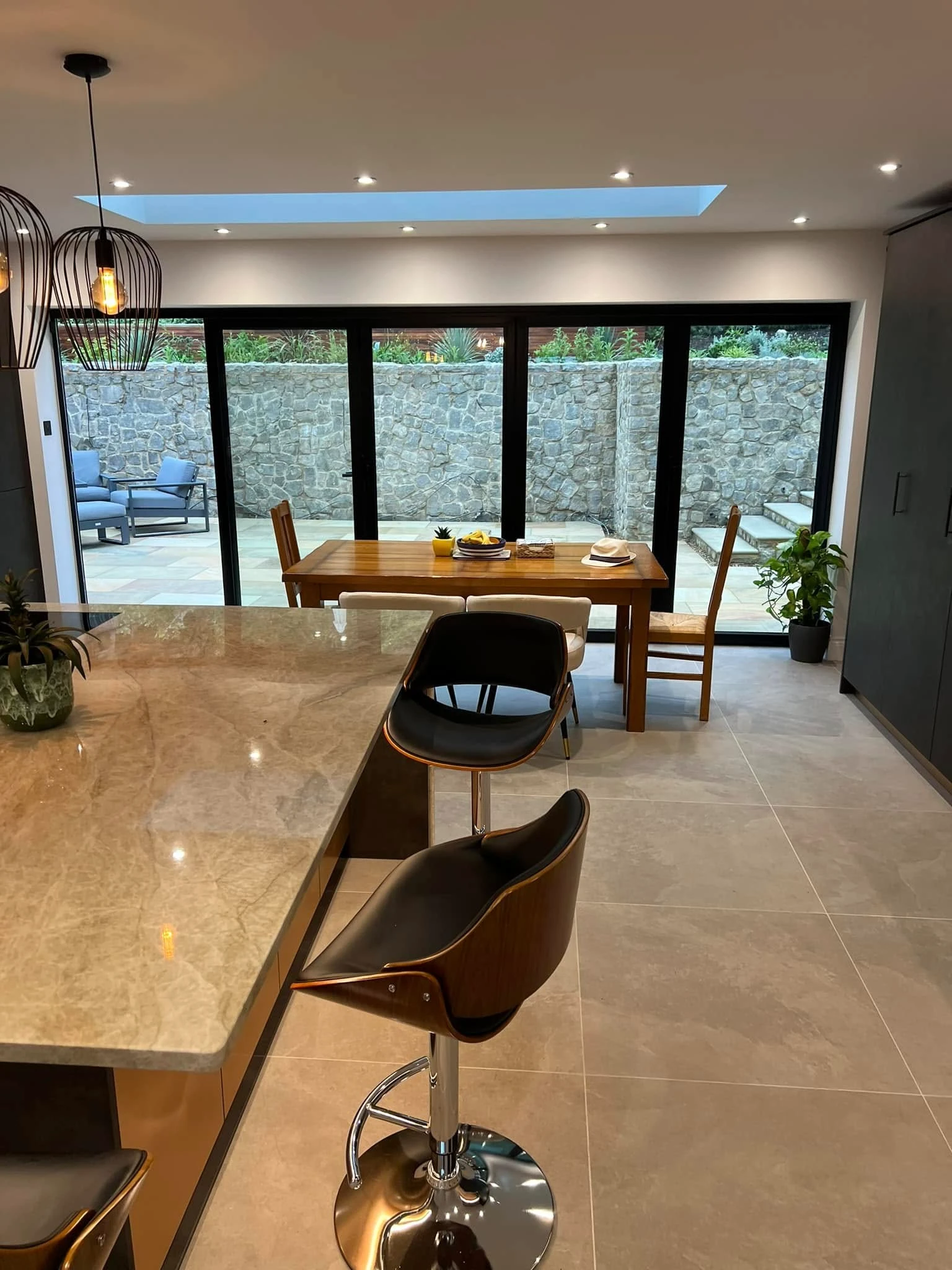 Modern kitchen with marble countertop island and two black bar stools, a wooden dining table with chairs, sliding glass doors showing an outdoor patio with stone wall and stairs.